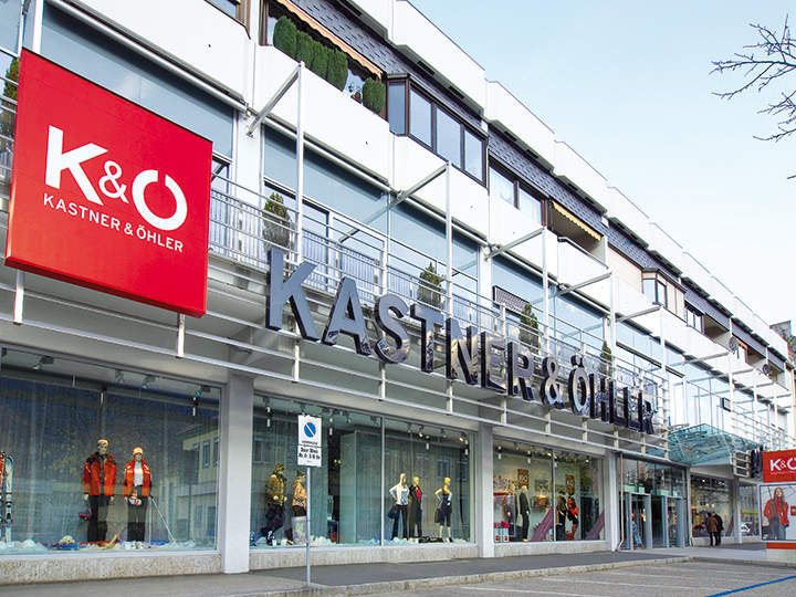 Das Bild zeigt die Ladenfront von Kastner & Öhler mit einem großen rot-weißen Logo auf der linken Seite und einer modernen Glasfassade. In den Fenstern unter dem markanten Namensschild des Ladens stehen Schaufensterpuppen in stilvoller Kleidung.