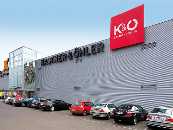 Ein großes Einkaufsgebäude mit einem roten Schild mit der Aufschrift „Kastner & Öhler“. Auf dem Parkplatz vor dem Gebäude parken mehrere Autos. Die Fassade ist metallisch grau, an der Seite verlaufen Fenster.