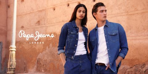 Eine Frau und ein Mann posieren vor einer strukturierten Steinwand. Beide tragen Jeanskleidung und weiße Hemden. Auf der linken Seite des Bildes ist das Logo von Pepe Jeans London zu sehen.
