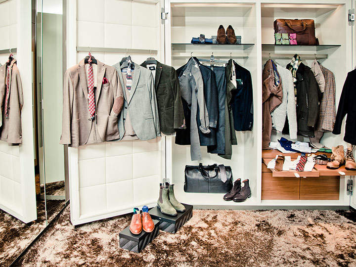 Eine stilvolle Herrengarderobe bietet eine Auswahl ordentlich hängender Anzüge und Jacken sowie elegante Schuhe, Stiefel und Accessoires wie Krawatten und Taschen. Der Schrank hat offene Regale und einen Spiegel an der Seite, gegenüber einem weichen Teppichboden.