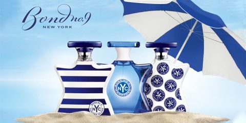 Drei Bond No. 9 New York-Parfümflaschen stehen auf Sand vor einem blauen Himmel. Die Flaschen sind mit Streifen und Mustern versehen. Im Hintergrund ist ein großer blau-weißer Sonnenschirm aufgespannt.