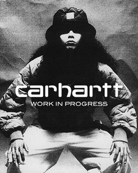 Schwarzweißbild einer Person mit Hut und Jacke, die mit den Händen auf den Knien hockt. Der Text „Carhartt Work In Progress“ ist deutlich in der Mitte zu sehen.