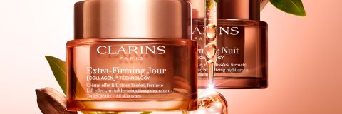Zwei Clarins-Hautpflegetiegel mit den Aufschriften „Extra-Firming Jour“ und „Extra-Firming Nuit“ hängen vor einem orangefarbenen Hintergrund mit Farbverlauf. Weiße Blüten und eine grüne Samenkapsel umspielen sie. Der Text hebt die Vorteile von Kollagen und eine um 53 % gesteigerte Festigkeit hervor.