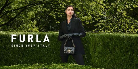 Eine Frau steht vor üppigem Grün. Sie trägt ein schwarzes Outfit und Handschuhe und hält eine schwarze gesteppte Handtasche in der Hand. Links ist der Text „Furla Since 1927 Italy“ zu sehen.