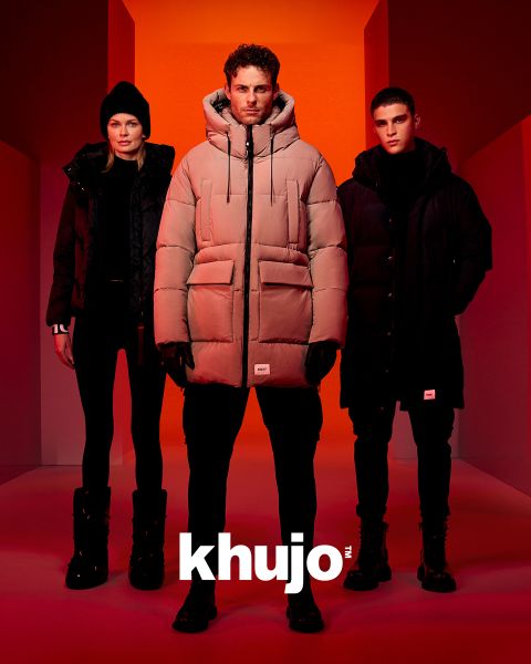 Drei Models stehen in einer Reihe, gekleidet in Winteroutfits mit Pufferjacken und Stiefeln, vor einem rot-orangefarbenen Hintergrund. Unten in der Mitte ist deutlich das Wort „khujo“ zu sehen.