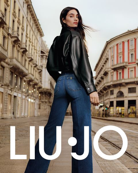 Eine Frau steht in einer Stadtstraße. Sie trägt eine schwarze Lederjacke und blaue Jeans. Sie blickt über ihre Schulter, während ihr langes Haar im Wind weht. Im Hintergrund ist klassische Architektur zu sehen. Unten steht der Text „LIU JO“.
