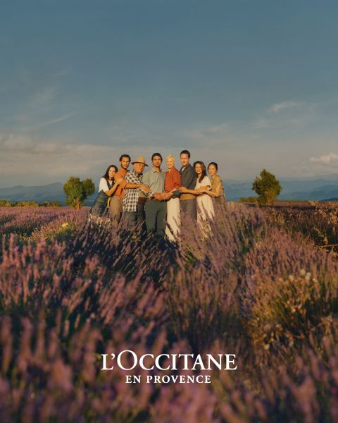 Eine Gruppe von Menschen steht zusammen in einem Lavendelfeld und hält sich an den Händen unter einem klaren Himmel. Sie lächeln in die Kamera, umgeben von leuchtend violetten Blumen. Unten ist der Markenname „L'OCCITANE EN PROVENCE“ zu sehen.