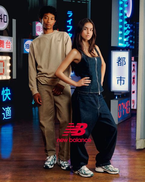 Ein Mann und eine Frau posieren vor Neonreklamen. Der Mann trägt einen beigen Trainingsanzug, die Frau einen Jeans-Overall. Beide tragen New Balance-Sneaker. Die Szene wird von verschiedenen Neonlichtern in Blau und Rot beleuchtet.