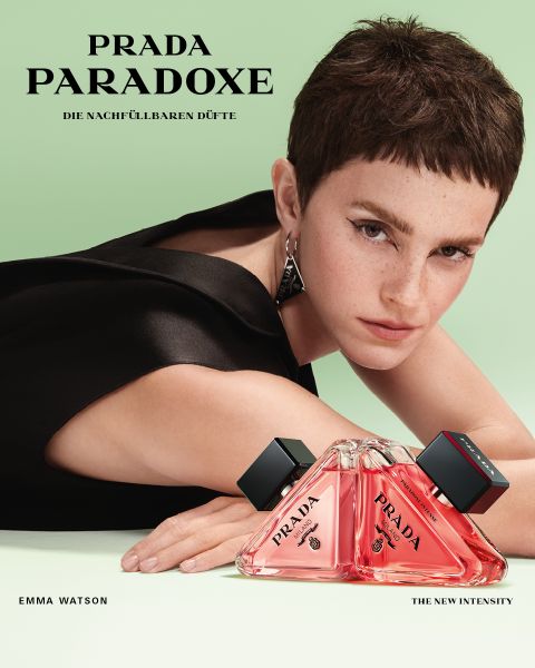 Eine Person mit kurzen Haaren und einem schwarzen Oberteil posiert neben einer dreieckigen Flasche Prada Paradoxe-Parfüm. Der grüne Hintergrund unterstreicht das elegante Design der Parfümflasche. Der Text enthält „Prada Paradoxe“ und „Emma Watson“.