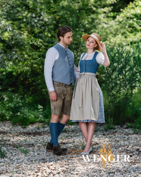Ein Mann und eine Frau stehen in traditioneller Kleidung auf felsigem Boden. Der Mann trägt eine blaue Weste, ein weißes Hemd, braune Shorts, blaue Socken und braune Schuhe. Die Frau trägt ein blaues Kleid mit einer weißen Bluse und einer Schürze sowie einen Strohhut. Sie sind von Grün umgeben.