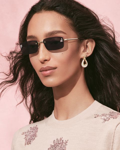 Eine Person mit welligem dunklem Haar, einer Sonnenbrille, einem cremefarbenen Oberteil mit floralen Verzierungen und hängenden Ohrringen posiert vor einem zartrosa Hintergrund.