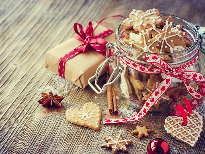 Eine festliche Szene mit einem Glas voller dekorierter Lebkuchen auf einem Holztisch. Daneben ein mit einer roten Schleife verpacktes Geschenk und Zimtstangen, die für festliche Stimmung sorgen. Weitere Kekse, Sternanis und ein rotes Ornament runden die Kulisse ab.
