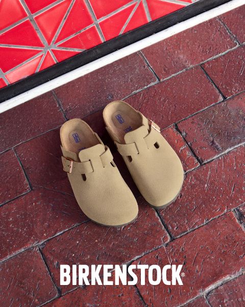 Ein Paar beigefarbene Birkenstock-Clogs mit Schnallenriemen auf einem roten Backsteinboden neben einer roten, geometrisch gefliesten Wand. Das weiße Birkenstock-Logo befindet sich am unteren Rand des Bildes.