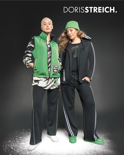 Zwei Frauen posieren selbstbewusst in schwarz-weißen Outfits mit grünen Akzenten, darunter eine Pufferweste, eine Mütze und Schuhe, vor einem dunklen Hintergrund mit dem Text "DORIS STREICH" über ihnen.