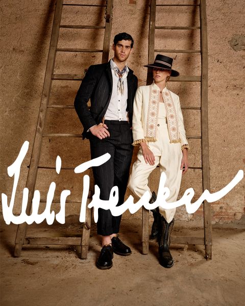 Ein Mann und eine Frau in stylischen, Vintage-inspirierten Outfits stehen zwischen zwei Holzleitern vor einer rustikalen Wand. Der Mann trägt einen dunkel gestreiften Anzug, die Frau einen cremefarbenen Anzug mit Hut und Stiefeln. „Luis Trenker“ steht in fetter Schrift darauf.