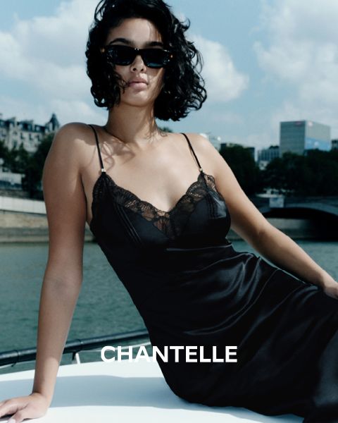 Eine Frau in einem schwarzen, spitzenbesetzten Slip-Kleid und mit Sonnenbrille sitzt auf einem Boot in der Nähe eines Stadtflusses, mit Gebäuden und Bäumen im Hintergrund. Das Wort "CHANTELLE" ist am unteren Rand des Bildes zu lesen.