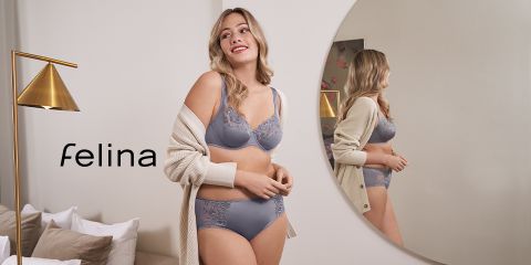 Eine Frau in grauer Unterwäsche steht lächelnd in einem Raum mit rundem Spiegel. Sie trägt eine beige Strickjacke und blickt vom Spiegel weg. Neben ihr steht eine moderne, goldene Stehlampe. Links ist das Wort „Felina“ zu sehen.