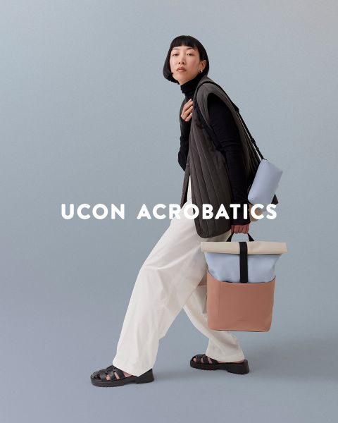 Eine Person mit Bob-Haarschnitt posiert vor einem gedeckten Hintergrund. Sie trägt ein schwarzes Oberteil, weiße Hosen und Sandalen und trägt einen stylischen Rucksack. Über dem Bild steht „Ucon Acrobatics“.