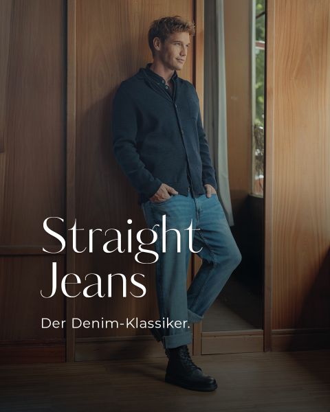 Ein Mann in einer marineblauen Strickjacke, blauen Straight-Jeans und schwarzen Stiefeln steht an einer Holzwand und einer Glastür. Der Text lautet: "Straight Jeans. Der Denim-Klassiker." Von draußen dringt natürliches Licht ein.