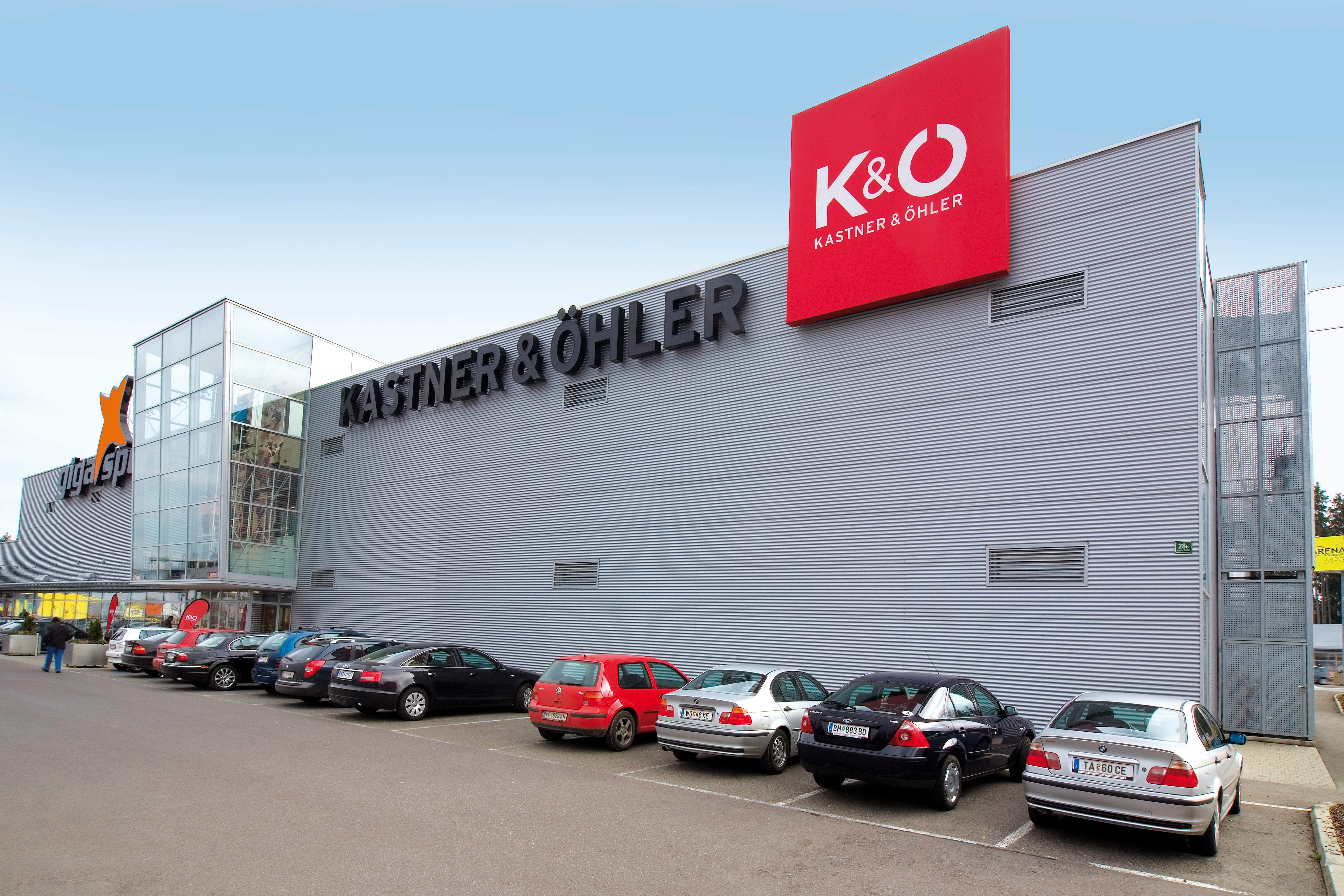 Ein modernes Geschäftshaus mit dem Schriftzug und Logo „Kastner & Öhler“ an der Fassade. Vor dem Gebäude, das auf der linken Seite eine große Glasfläche besitzt, parken mehrere Autos auf dem Parkplatz. Der Himmel ist klar.