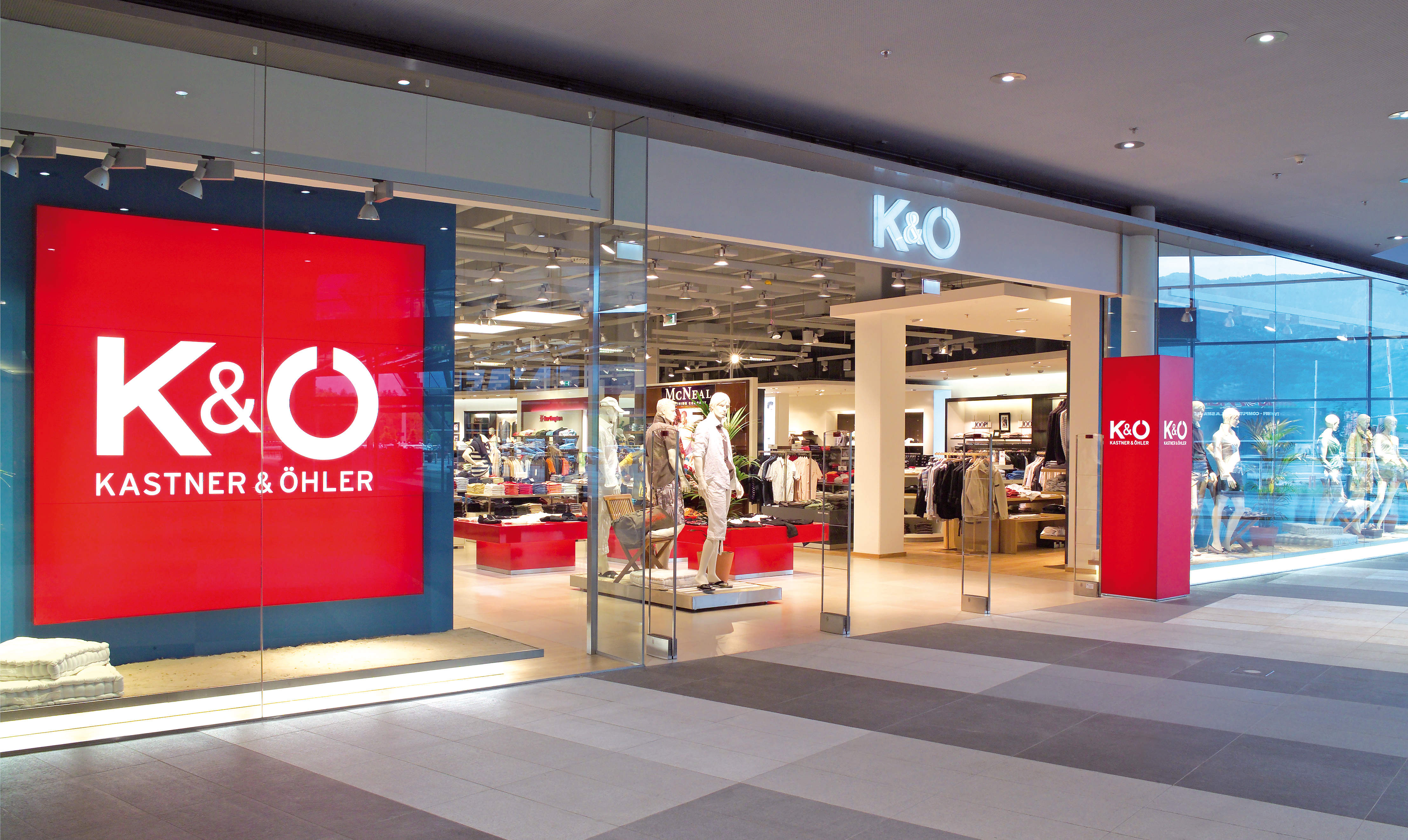 Ein moderner Ladeneingang mit großen Glasfenstern und -türen. Auf dem Ladenschild steht „K&Ö Kastner & Öhler“ und es sind Schaufensterpuppen in verschiedenen Outfits zu sehen. Der Innenraum ist gut beleuchtet und präsentiert Kleidung und Accessoires.