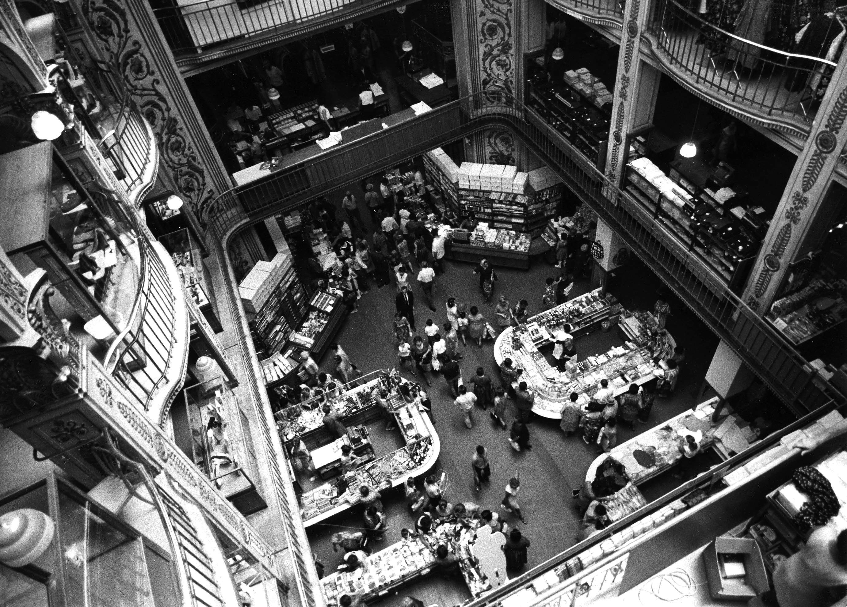 Eine schwarz-weiße Luftaufnahme eines überfüllten, mehrstöckigen Einkaufszentrums. Menschen gehen umher und stöbern an verschiedenen Ständen mit Waren und Büchern. Die Architektur zeichnet sich durch kunstvolle Details und geschwungene Balkone auf jeder Etage aus.