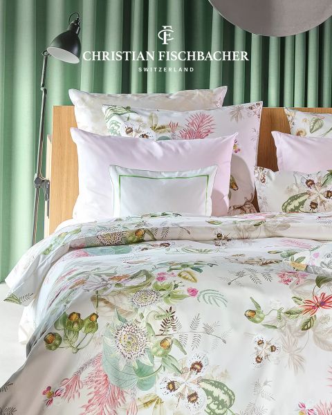 Ein ordentlich gemachtes Bett mit blumengemustertem Bettzeug und weißen Kissen steht vor einem grünen Vorhang. Das Logo von Christian Fischbacher Switzerland erscheint oben auf dem Bild.