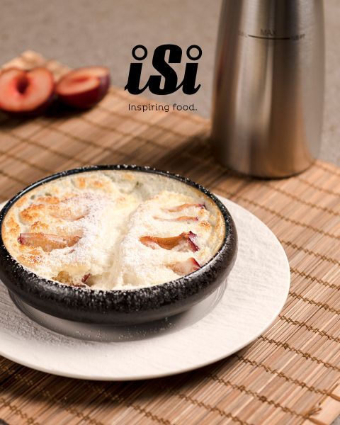 Ein mit Puderzucker bestäubtes ofengebackenes Dessert wird in einer schwarzen Schüssel serviert, im Hintergrund sind Pflaumen und ein Spender aus Metall zu sehen.
