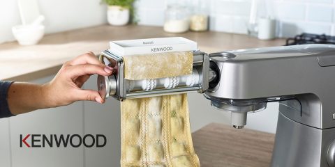 Eine Person verwendet eine Kenwood-Küchenmaschine mit Nudelaufsatz, um Ravioli zuzubereiten. Der frische Nudelteig wird ausgerollt und in Raviolistücke geschnitten. Die Küche ist modern eingerichtet, mit Gläsern und einer Kaffeetasse im Hintergrund.