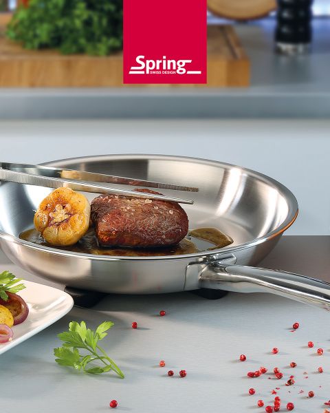Auf einer Küchenarbeitsplatte steht eine Edelstahlpfanne mit einem Steak und Knoblauch, das mit einer Zange gewendet wird. Daneben liegen rote Pfefferkörner und Petersilie.
