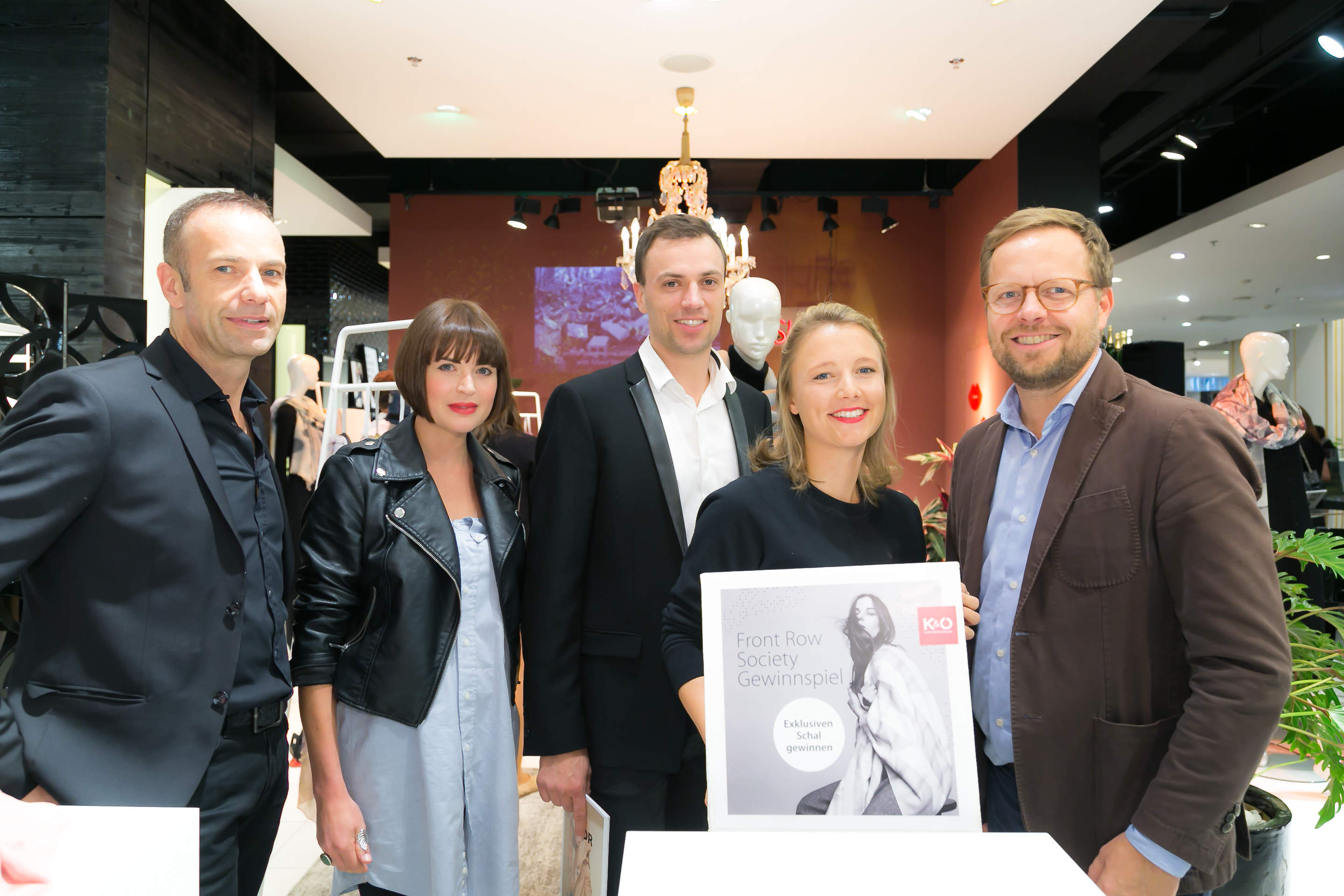 Fünf Personen stehen bei einer Indoor-Veranstaltung zusammen und lächeln. Sie sind in Business-Casual-Kleidung gekleidet. Zwei von ihnen halten ein Plakat mit dem Text „Front Row Society Gewinnspiel“ in der Hand. Im Hintergrund sind eine rote Wand und Schaufensterpuppen zu sehen.