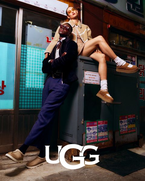 Zwei Models posieren in einer urbanen Umgebung und präsentieren stylische UGG-Schuhe. Der Mann lehnt lässig in blauem Outfit an einer Wand, während die Frau in einem hellbraunen Outfit über ihm sitzt. Beide tragen beige UGG-Schuhe. Unten ist das UGG-Logo zu sehen.