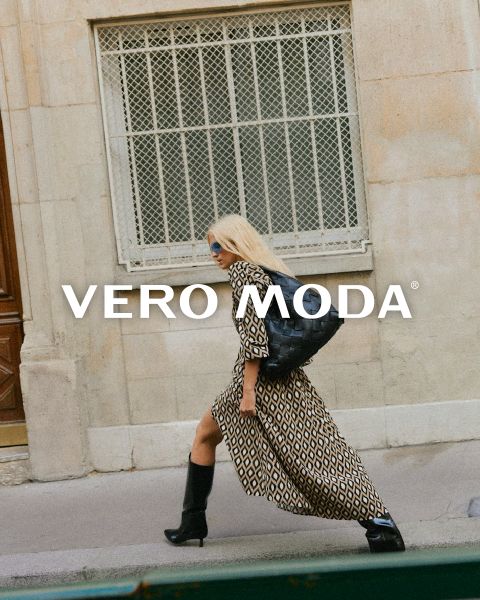 Frau in geometrisch gemustertem Kleid und schwarzen Stiefeln geht an einem Gebäude mit vergitterten Fenstern vorbei. Sie trägt einen schwarzen Rucksack und eine Sonnenbrille. Auf dem Bild ist das VERO MODA-Logo zu sehen.