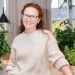 Eine lächelnde Frau mit Brille und roten Haaren steht in einem gemütlichen Raum mit großen Fenstern. Sie trägt einen beigen Pullover und ist von üppigen grünen Pflanzen umgeben. Natürliches Licht erhellt den Raum und schafft eine warme und einladende Atmosphäre.