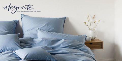 Ein ordentlich gemachtes Bett mit weicher blauer Bettwäsche steht vor einer weißen Wand. Auf einem kleinen Holzregal rechts steht eine weiße Vase mit getrockneten Blumen. In der oberen linken Ecke befinden sich ein elegantes Logo und ein Text.