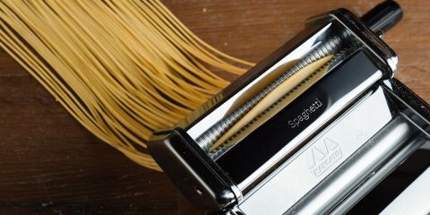 Eine Nudelmaschine presst Spaghetti auf eine Holzoberfläche. Die Maschine ist aus glänzendem Metall gefertigt, auf der Walze ist ein Etikett zu sehen. Die Nudelstränge kommen ordentlich ausgerichtet aus der Maschine.