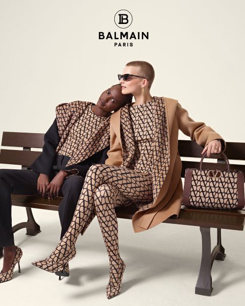 Zwei Models sitzen in passenden Balmain-Outfits mit geometrischen Mustern auf einer Bank. Eines lehnt sich an das andere und trägt ein schwarzes Top und eine Hose, während das andere einen Kamelhaarmantel und eine Sonnenbrille trägt. Neben ihnen steht eine gemusterte Handtasche. Oben das Balmain-Logo.