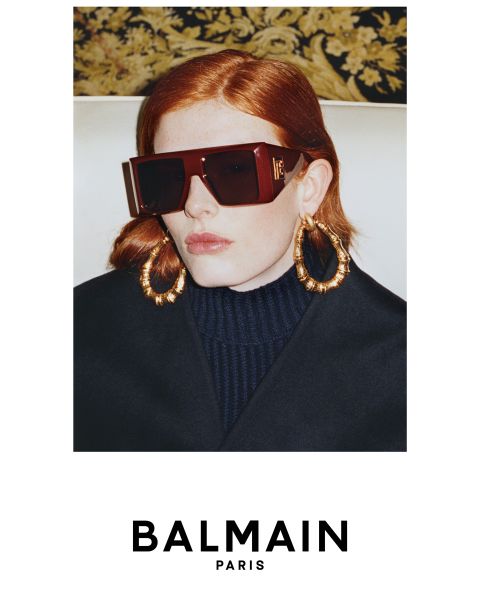 Eine Person mit roten Haaren, einer übergroßen dunklen Sonnenbrille, großen goldenen Ohrringen und einem dunklen hochgeschlossenen Oberteil posiert vor einem gemusterten Hintergrund. Das Bild zeigt das BALMAIN Paris Logo am unteren Rand.