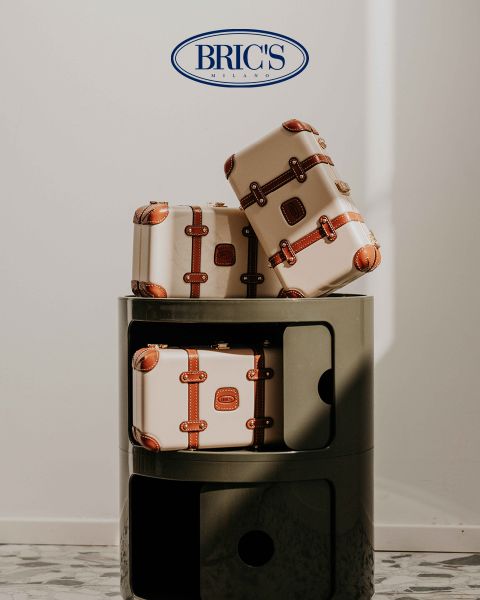 Drei beige Koffer mit braunen Lederriemen stapeln sich auf einem modernen, runden, dunkelgrauen Regal. Darüber prangt das BRIC'S MILANO-Logo auf einer weißen Wand. Sonnenlicht wirft sanfte Schatten auf die Szene.