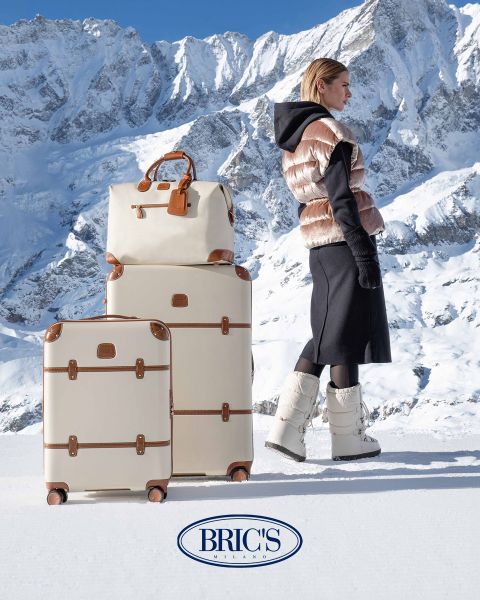 Eine Person in Winterkleidung steht mit drei beigen und braunen Gepäckstücken auf Schnee. Den Hintergrund bilden schneebedeckte Berge. Unten ist das Logo „Bric’s Milano“ zu sehen.