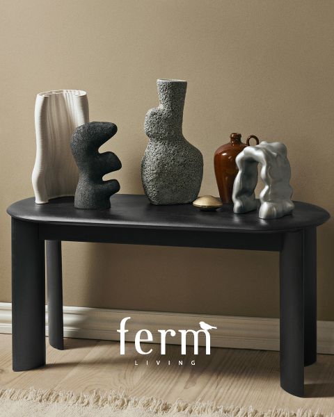 Ein schwarzer Tisch mit abstrakten Keramikskulpturen in verschiedenen Formen und Texturen steht vor einer beigen Wand. Der Boden besteht aus hellem Holz und unten ist das Logo „ferm LIVING“ zu sehen.