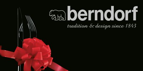 Ein Besteckset, bestehend aus Gabel, Messer und Löffel, ist mit einer roten Schleife zusammengebunden. Der Text lautet „berndorf tradition & design since 1843“, mit einem stilisierten Bärenlogo über „berndorf“. Der Hintergrund ist schwarz.