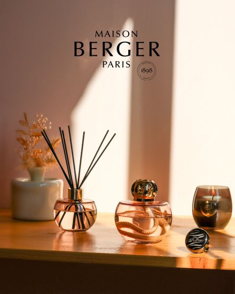 Auf einem sonnenbeschienenen Regal stehen zwei Diffusoren von Maison Berger Paris mit elegantem Glasdesign. Ein Diffusor ist mit Schilfrohren ausgestattet, der andere mit einer dekorativen Kappe. Daneben steht eine kleine Vase mit Trockenblumen und einer Kerze, die eine warme, einladende Atmosphäre schafft.
