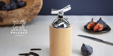 Eine Pfeffermühle aus Holz und Edelstahl steht neben ganzen und geschnittenen Feigen auf einer Tischdecke. Daneben liegt ein Messer. In der oberen linken Ecke ist das Peugeot-Logo mit einem Löwen zu sehen. Im Hintergrund ist eine Holzschale mit Gewürzkapseln zu sehen.