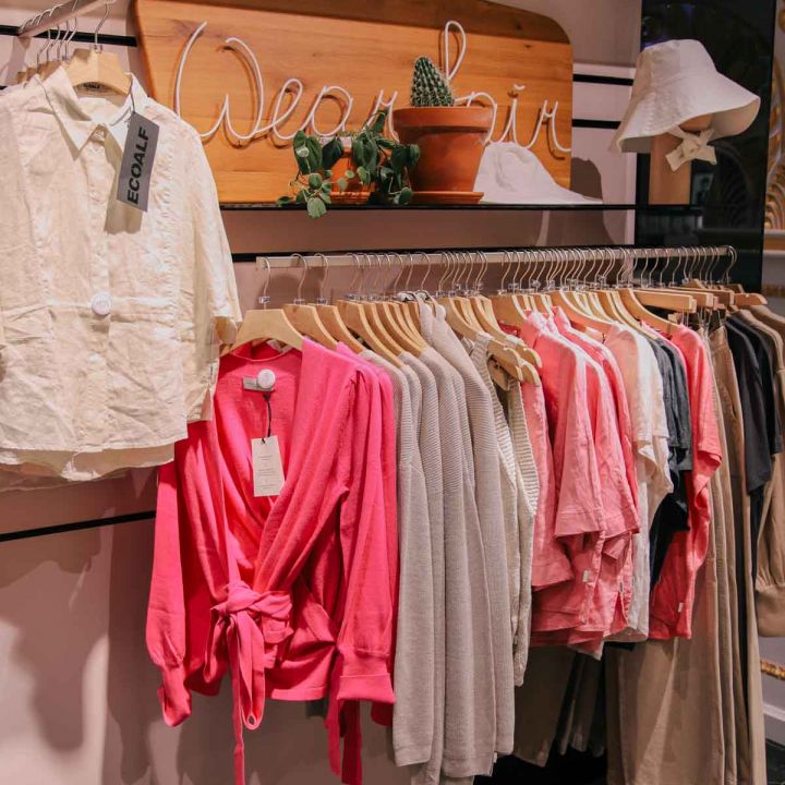 Auf einem Kleiderständer hängen verschiedene Kleidungsstücke, darunter ein weißes Hemd, eine rosa Strickjacke und mehrere Oberteile in neutralen Farben auf hölzernen Kleiderbügeln. Über dem Ständer hängt ein Holzschild mit Pflanzen. In der oberen rechten Ecke hängt ein weißer Hut.