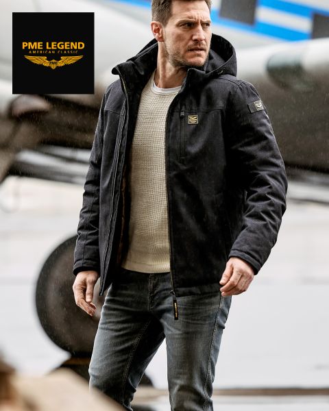 Ein Mann, der eine schwarze Kapuzenjacke, einen weißen Pullover und Jeans trägt, steht im Freien in der Nähe eines Flugzeugs. Das PME Legend American Classic-Logo erscheint in der oberen linken Ecke.