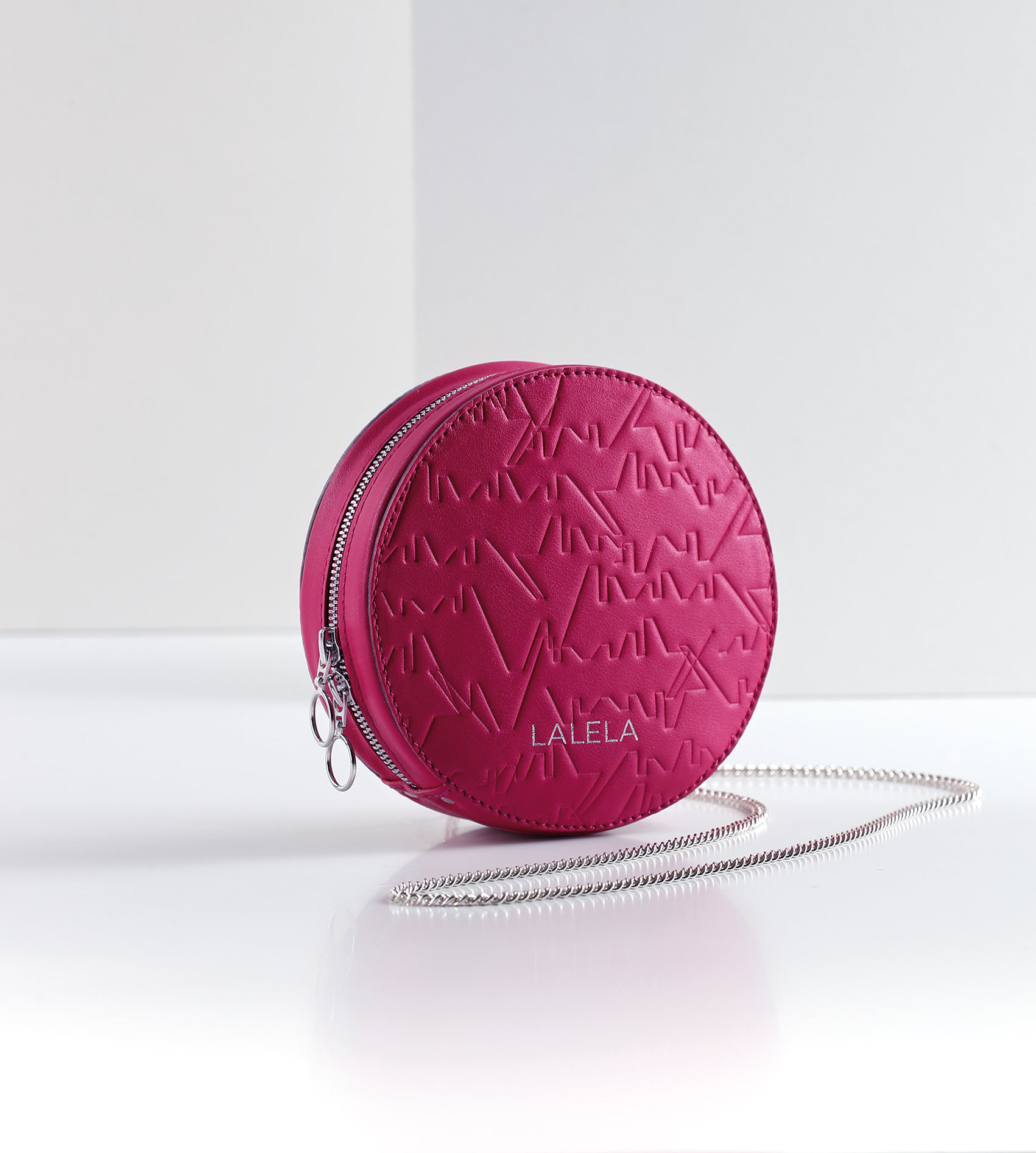 Eine runde, leuchtend rosa Handtasche mit Steppmuster und silbernem Kettenriemen liegt auf einer weißen Oberfläche. Auf der Vorderseite ist das Wort „LALELA“ eingeprägt. Der Hintergrund ist in schlichtem Hellgrau gehalten, was für einen minimalistischen Look sorgt.