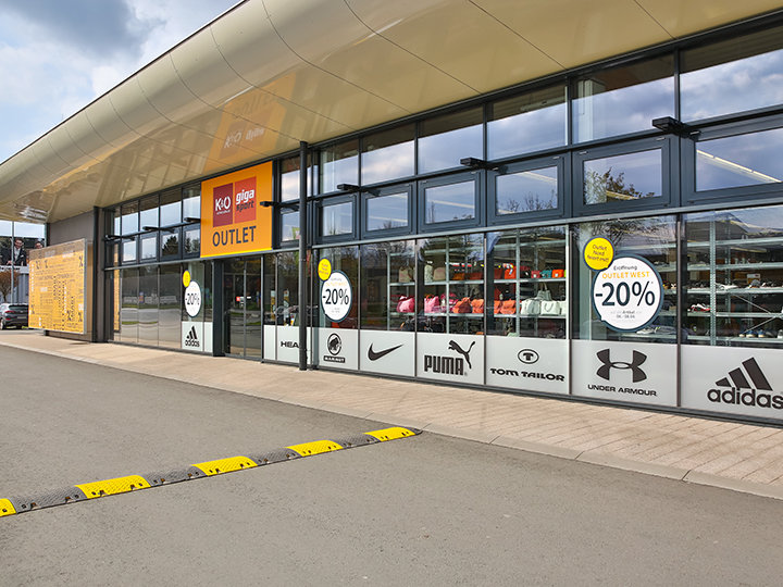 Außenansicht eines Outlet-Stores mit Glasfenstern, auf denen Markenlogos von Puma, Adidas und Under Armour zu sehen sind. Schilder werben mit 20 % Rabatt. Das Gebäude hat ein gelbes Vordach und davor einen Fußgängerweg.