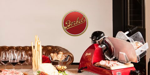 Auf einem Tisch mit verschiedenen Fleischsorten, Käse, Grissini und Weingläsern steht eine alte Aufschnittmaschine für rotes Fleisch. Im Hintergrund sind ein Ledersofa und ein „Berkel“-Logo zu sehen. Das Ambiente ist gemütlich und rustikal.