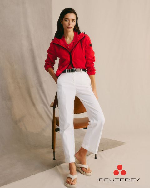Eine Frau in roter Jacke und weißer Hose lehnt an einem Stuhl. Sie hat langes dunkles Haar und trägt weiße Sandalen. Die Kulisse ist minimalistisch mit neutralem Hintergrund. Das „Peuterey“-Logo befindet sich in der unteren rechten Ecke.